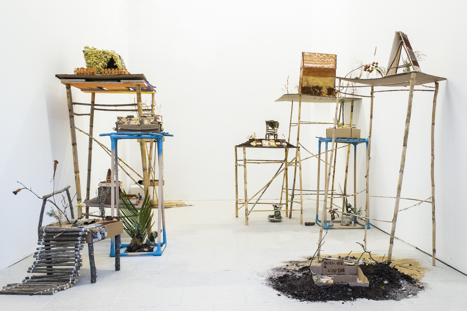 Photographie d'une salle d'exposition et de plusieurs constructions sur pilotis en cannes de Provence, sur lesquelles sont présentées des paysages et des constructions miniatures fabriqués à partir de matériaux récupérés ou naturels.