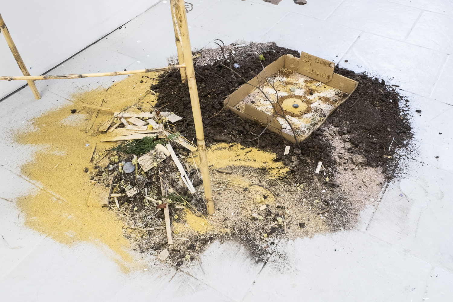 Photographie en plongé et d'un paysage miniature posé au sol et fabriqué à partir de matériaux récupérés, naturels et de balayures, où il est marqué sur une pancarte avec de la terre « du chaos à l'ordre ».