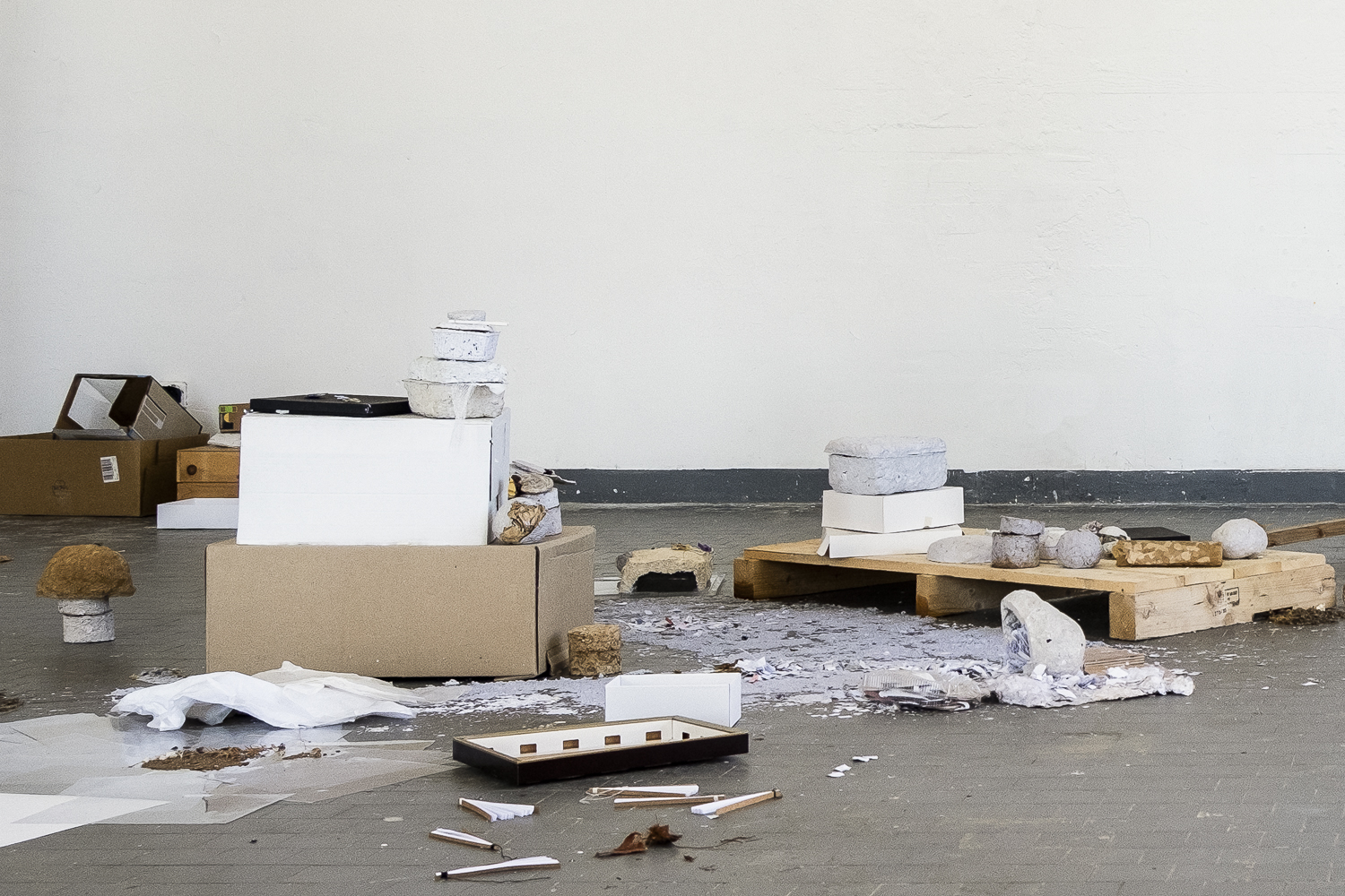 Photographie d'une salle d'exposition où une installation avec du papier déchiqueté, mixé et mouillé, des objets blancs ou marrons et fabriqués en papiers collés ou mâchés, ainsi que des balayures sont présentés au sol, sur des planches et sur des cartons.