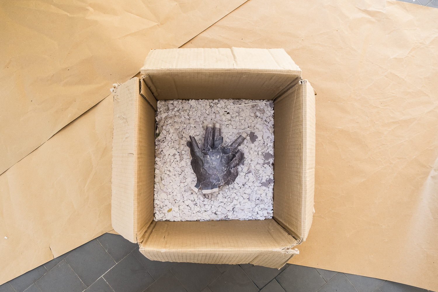 Photographie en plongée d'un carton posé au sol sur des feuilles de papiers krafts. À l'intérieur du carton est posé le moulage en papier avec une impression violette d'une main posée sur un tapis de papiers blancs et violets déchiquetés.