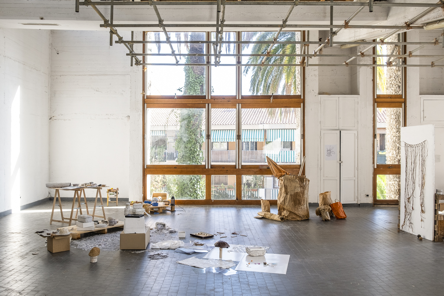 Photographie d'une salle d'exposition où des objets blancs ou marrons et fabriqués en papiers collés ou mâchés sont présentés au sol, sur des planches, sur des cartons et sur une table à tréteaux.