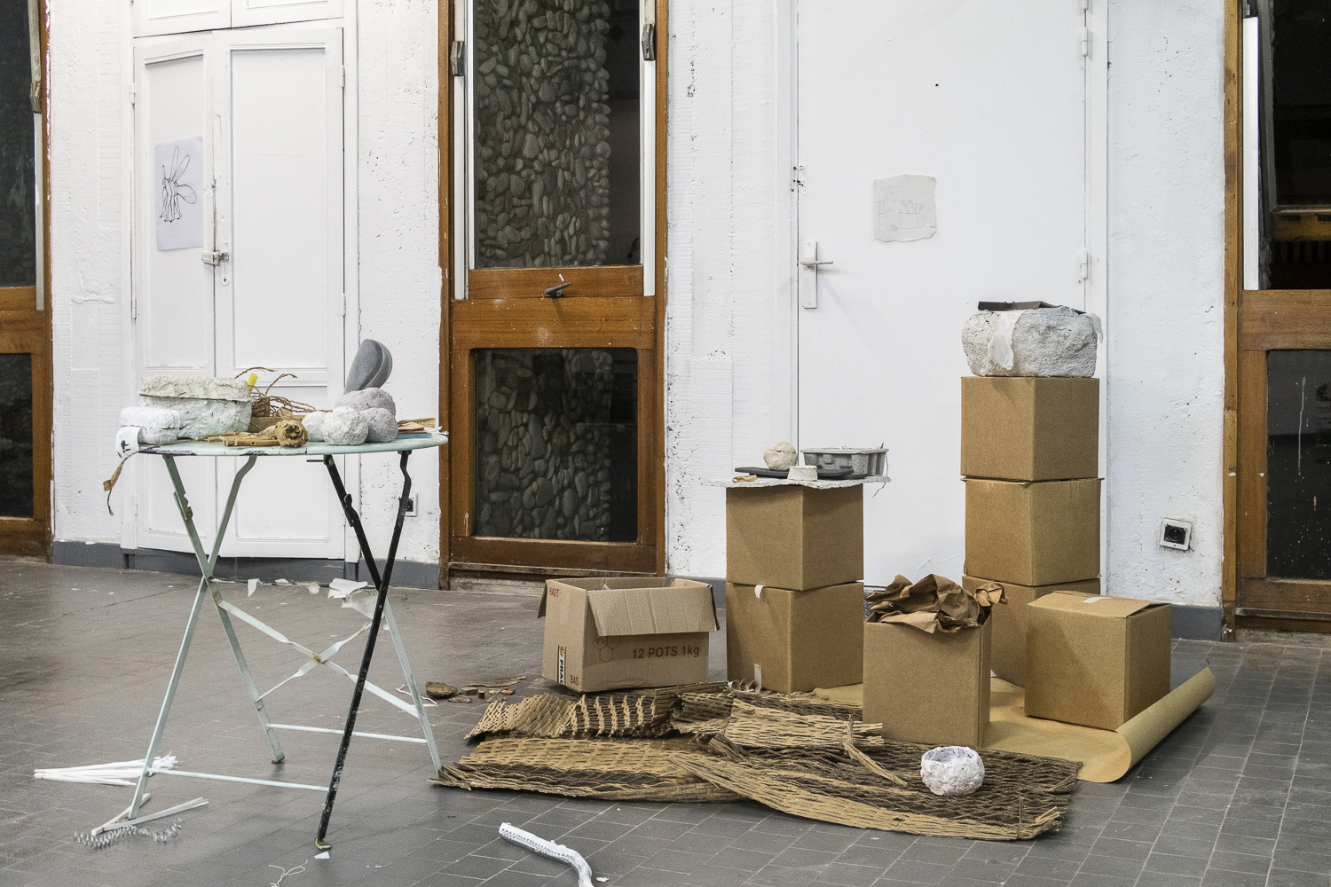 Photographie d'une salle d'exposition, où des papiers, des objets en plastiques et des sculptures en papier mâché sont présentés sur une table en fer ronde et des cartons empilés comme socles.