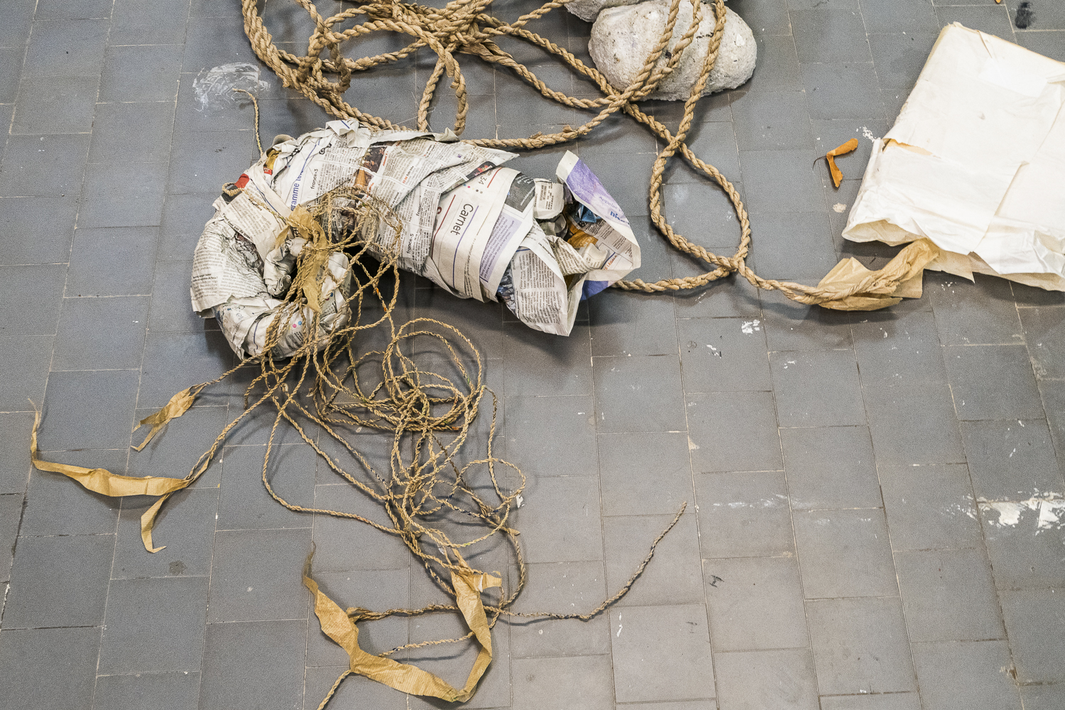 Photographie d'une salle d'exposition où des graines ainsi que des objets fabriqués en papiers sont posés au sol, dont des cordes et des ficelles en papier kraft et une grande spirale en papier journal.