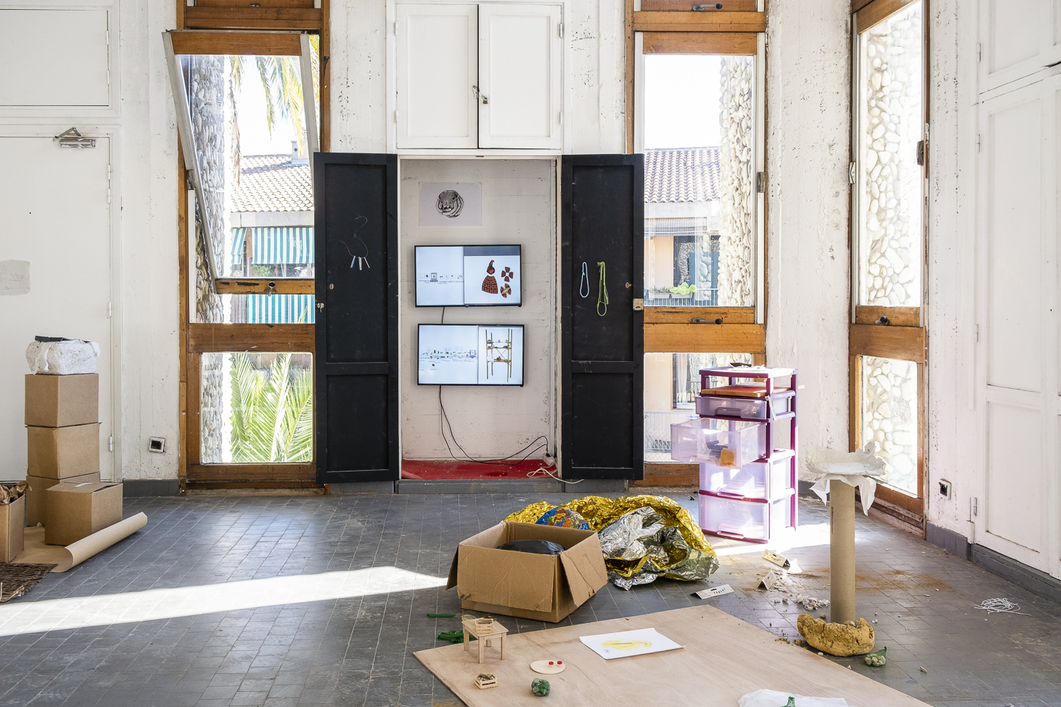 Photographie d'une salle d'exposition où deux écrans diffusent des vidéos dans un placard et où des objets comme une couverture de survie, des tiroirs en plastiques, des cartons et de petits emballages sont posés au sol, ainsi qu'une table et des cagettes en bois miniatures.