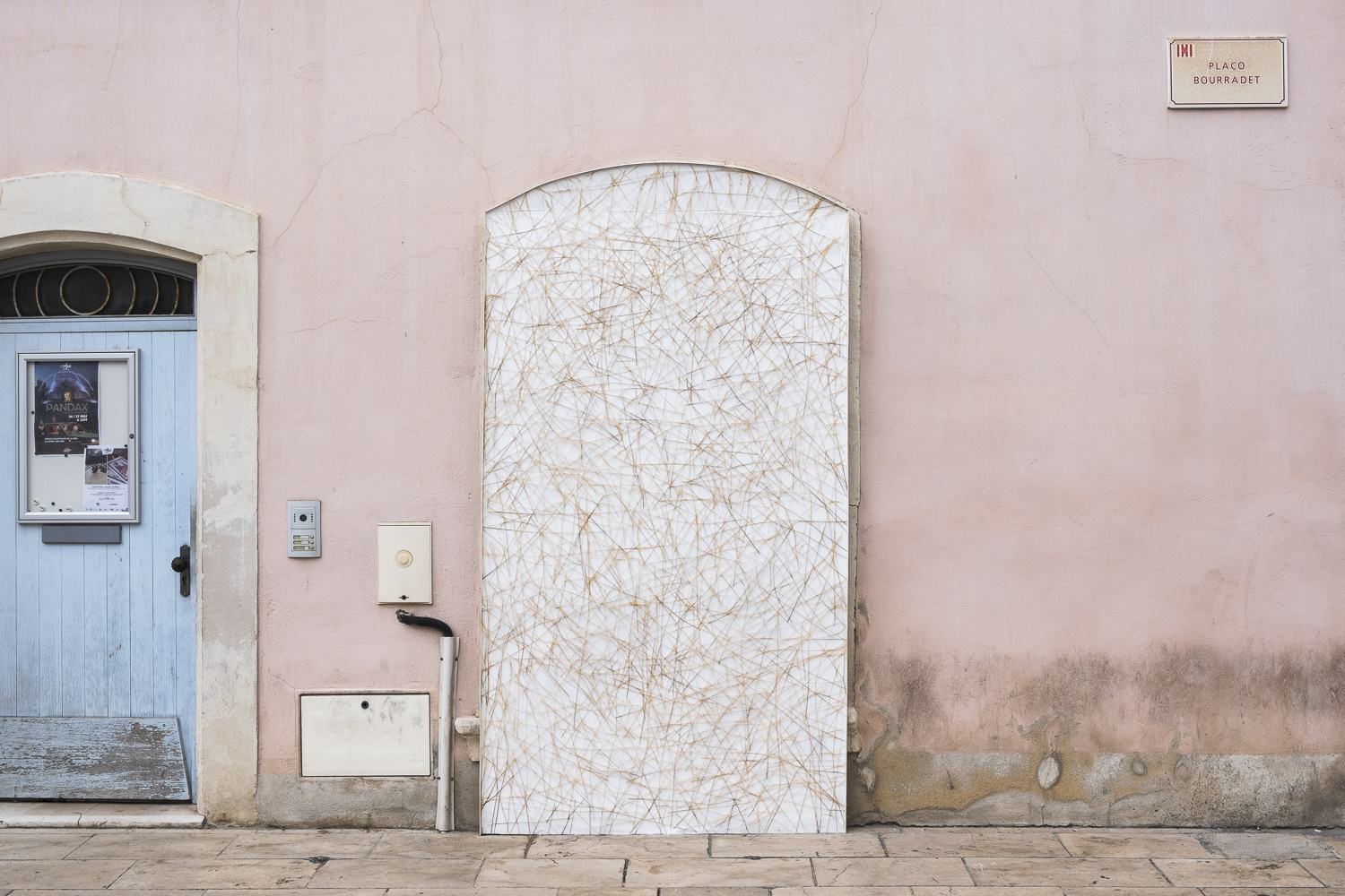 Photographie d'un support blanc contre une façade en forme de porte, où des graminées sont entrelacées et fixées.